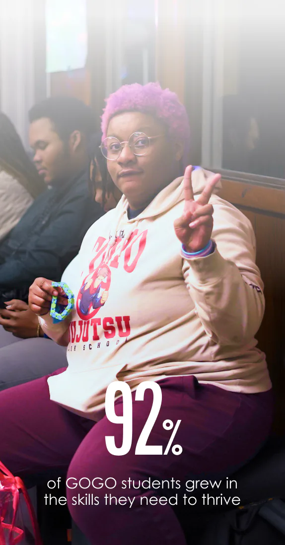 Person with pink hair and glasses wearing a light-colored hoodie with a cartoon character on it shows a peace sign while sitting on a bench, with two other people seated nearby. Text at the bottom reads: 92% of GOGO students grew in the skills they need to thrive.
